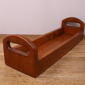 May include: A rectangular wooden tray with a medium brown finish, featuring two raised end panels with integrated oval cut-out handles. The tray has a flat base and low side walls, showcasing a visible wood grain pattern throughout the structure.