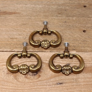 May include: Three antique brass drawer pulls with ornate, scrolled designs. Each pull has a ring shape with a decorative center and a screw for mounting. The pulls are set against a light brown wooden surface.