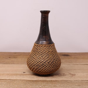May include: A brown ceramic vase with a woven brown wicker exterior. The vase has a narrow neck and a rounded base.
