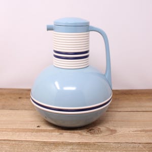 May include: A light blue vintage thermos with a white and navy striped neck and a matching lid. The thermos has a rounded base and a curved handle. It is sitting on a wooden surface.