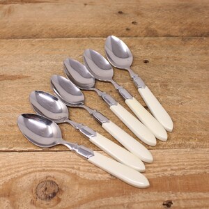 May include: Six silver spoons with white handles. The spoons are arranged in a row, with the handles pointing to the right. The spoons are on a wooden surface.