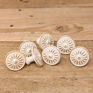 May include: Set of six white ceramic cabinet knobs with a gold rim and a raised sunburst design.