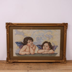 May include: A framed needlepoint artwork depicting two cherubs with wings, looking up at the sky. The cherubs are rendered in shades of pink, blue, and yellow against a light blue background. The frame is gold with ornate detailing.