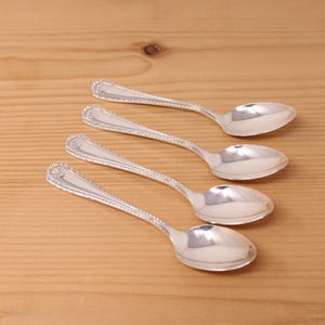 May include: A set of four polished silver-toned spoons arranged diagonally. Each spoon features a slender handle with a decorative beaded edge and a small shell-like motif at the top of the handle, leading to a smooth, oval-shaped bowl.