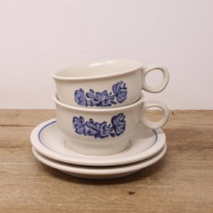 May include: A set of three white ceramic teacups with blue floral designs. The cups are stacked on top of each other and are sitting on a wooden surface.