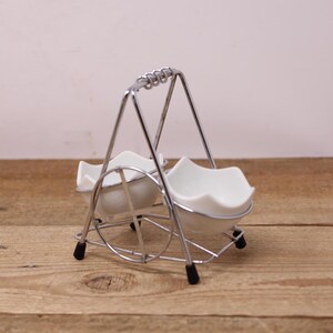 May include: A vintage egg holder with two white porcelain egg cups. The cups have a wavy rim and are held in a chrome-plated wire frame with a handle. The holder has black rubber feet and is set on a wooden surface.