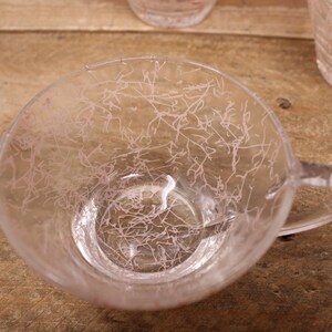 May include: A clear glass cup with a pink design. The cup has a handle and is sitting on a wooden surface.
