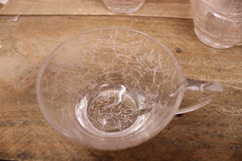 May include: A clear glass cup with a pink design. The cup has a handle and is sitting on a wooden surface.