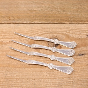 May include: Four silver cocktail forks with ornate handles. The forks are arranged in a row on a wooden surface.