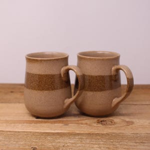 Peut inclure: Deux tasses en céramique avec une finition beige clair mouchetée et une large bande horizontale de glaçure brun foncé autour du centre. Chaque tasse présente un corps arrondi, un bord lisse et une anse incurvée fixée sur le côté.