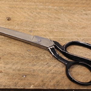 May include: Vintage scissors with a silver blade and black handles. The scissors are on a wooden surface. The handles show signs of wear. The blades are approximately 15 cm long.