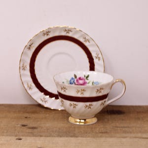 May include: A white porcelain teacup and saucer set featuring a deep maroon band, gold trim, and small gold leaf accents. The teacup displays a floral pattern with a pink rose and blue flowers, while both pieces have a fluted, scalloped edge design.