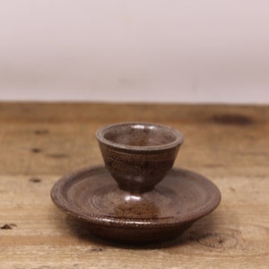 May include: A brown ceramic egg cup with a matching saucer. The cup has a slightly flared rim and a rounded base. The saucer is slightly wider than the cup and has a slightly raised edge.