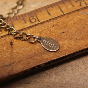 May include: A gold chain necklace with a small, round, gold charm that says "Alice + Eve". The necklace is lying on a wooden ruler.