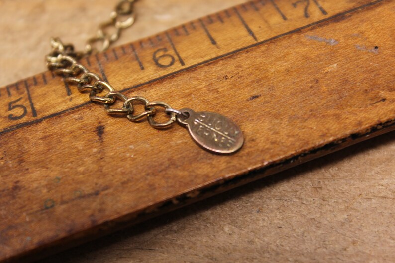 May include: A gold chain necklace with a small, round, gold charm that says "Alice + Eve". The necklace is lying on a wooden ruler.