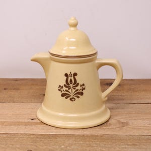 May include: A yellow ceramic creamer with a brown floral design and a lid. The creamer has a rounded body and a spout on one side. The lid has a knob on top.