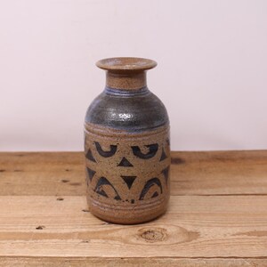 Peut inclure: Un vase en céramique fait à la main avec un corps cylindrique et un dessus évasé. Le vase présente une base beige mouchetée, un dessus bleu foncé et un motif répétitif de formes géométriques noires. Le vase est posé sur une surface en bois.