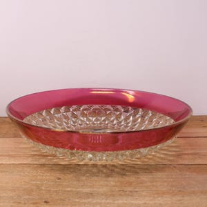 May include: A clear glass serving bowl with a ruby red rim. The bowl features a diamond-cut pattern in the center. The rim is a vibrant, glossy red, adding a pop of colour to the clear glass. It sits on a wooden surface.