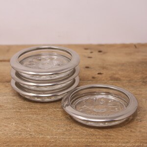 Peut inclure: Un ensemble de six sous-verres en verre avec des bords argentés. Les sous-verres sont empilés les uns sur les autres, avec un sous-verre posé à plat sur une surface en bois.