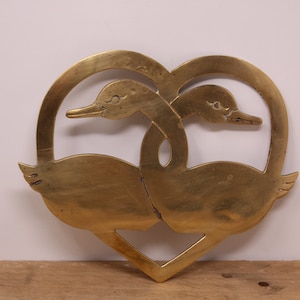 May include: A brass heart-shaped ornament featuring two swans intertwined. The swans are facing each other with their necks curved around each other.