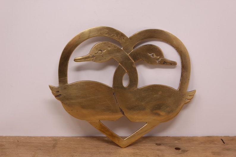 May include: A brass heart-shaped ornament featuring two swans intertwined. The swans are facing each other with their necks curved around each other.
