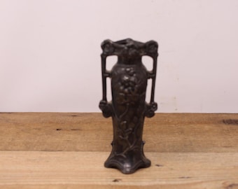 May include: A small, silver-coloured metal vase with an ornate design featuring floral motifs. The vase has a narrow neck and a wide base.