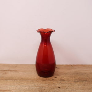 Peut inclure: Un vase en verre rouge rubis avec un bord festonné. Le vase a un corps élancé et allongé et repose sur une surface en bois. Le vase est vide et a une finition brillante.
