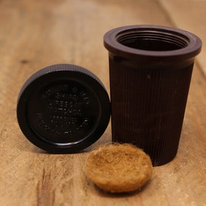 May include: A brown, cylindrical container with a matching lid and a small, round, fibrous object. The lid has embossed text. The container has vertical ridges. The items are on a wooden surface.