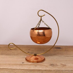Peut inclure: Un pot de fleurs suspendu en cuivre avec un support et une chaîne en laiton. Le pot est rond et a une finition brillante.