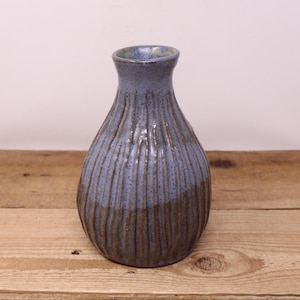 May include: A handmade ceramic vase in blue and brown. The vase has a textured, ribbed design and a rounded body that tapers to a narrow neck. The vase is displayed on a wooden surface.