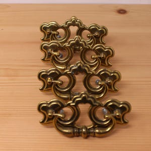 May include: A set of four brass drawer pulls featuring an ornate, symmetrical design with a central arched top and curved, swinging handles. Each pull has a polished metallic finish with darkened recessed areas that highlight the intricate details.