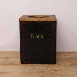 May include: A vintage wooden flour canister with a black painted body and a red and white border. The lid is made of wood and has a knob on top. The word "Flour" is written in gold lettering on the front of the canister.