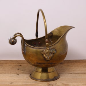 May include: A brass coal scuttle with a porcelain handle and a decorative design. The scuttle has a handle that can be used to carry it and a base to stand on.