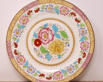 May include: A round, decorative plate with a floral design. The plate features a cream-colored background with a central arrangement of pink, red, and yellow flowers. The border has a repeating floral pattern in pink and blue, with a gold trim.