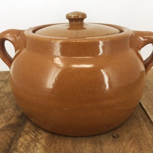 May include: A brown ceramic bean pot with a lid and two handles. The pot is glazed and has a rustic look.
