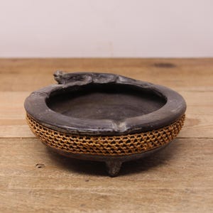 May include: A dark grey ceramic bowl with a woven, light brown mesh band around the base. The bowl has three small feet and a small, decorative element on the rim. The bowl is sitting on a wooden surface.