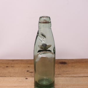 May include: A light green glass bottle with a distinctively shaped neck featuring a small internal glass marble. The bottle has a cylindrical body with a smooth, translucent finish and a narrow opening at the top, showcasing a classic design.