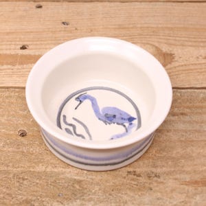 May include: A small, white ceramic bowl with a blue heron design. The bowl has a rounded shape with a wide rim and a blue and gray band around the base. The heron is depicted in blue within a circular frame.