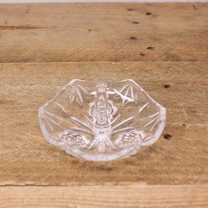 May include: A clear glass ring dish with a decorative design and a handle. The dish is on a wooden surface.