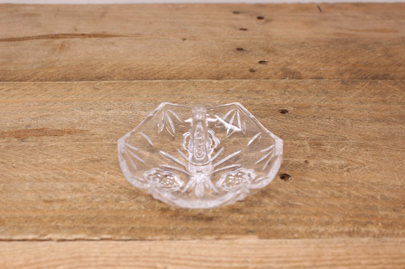 May include: A clear glass ring dish with a decorative design and a handle. The dish is on a wooden surface.