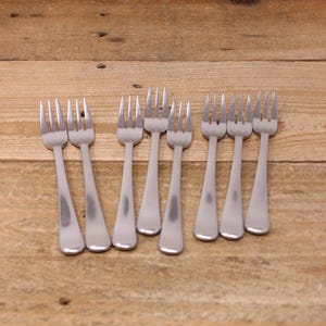 May include: Eight silver forks laid out on a wooden surface. The forks are all the same size and shape, with a simple design.