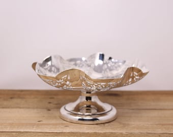 May include: A silver, decorative, footed bowl with an openwork design. The bowl is on a wooden surface.