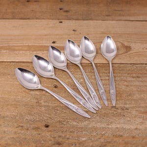 Peut inclure: Six cuillères en argent disposées sur une surface en bois. Les cuillères ont un design classique avec un bol arrondi et un manche fin. Les manches sont décorés d'un motif floral. Les cuillères sont réfléchissantes, et la surface en bois a un grain naturel.