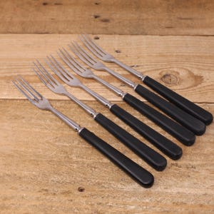 May include: Six stainless steel forks with black handles are arranged diagonally on a wooden surface. The forks have a classic design, with tines and a polished metal finish contrasting with the matte black handles.