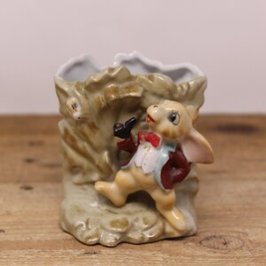 Peut inclure: Un pot en céramique en forme de falaise rocheuse avec une figurine de lapin debout sur le dessus. Le lapin porte une veste rouge, une chemise bleue et un nœud papillon. Le pot est peint dans des tons de brun et de vert.