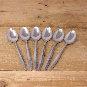 May include: Six stainless steel spoons with a decorative pattern on the handles. The spoons are arranged on a wooden surface, showcasing their polished, reflective surfaces. The spoons are designed for eating.