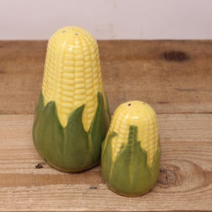 May include: A set of two ceramic salt and pepper shakers shaped like ears of corn. The shakers are yellow and green with brown details.