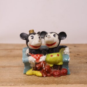 May include: A vintage figurine of Mickey Mouse and Minnie Mouse sitting on a blue couch. Mickey is wearing a red and white polka dot shirt and Minnie is wearing a green and yellow shirt with red polka dots.