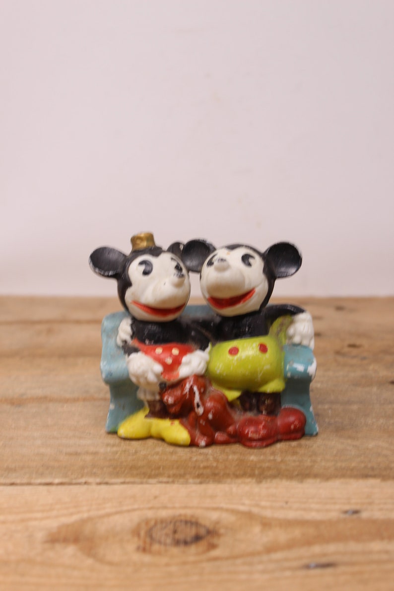 May include: A vintage figurine of Mickey Mouse and Minnie Mouse sitting on a blue couch. Mickey is wearing a red and white polka dot shirt and Minnie is wearing a green and yellow shirt with red polka dots.