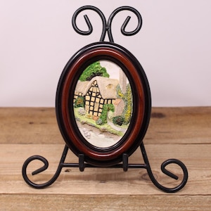May include: An oval-shaped decorative frame with a miniature cottage scene. The frame has a dark wood-tone border and a black metal stand. The cottage features a thatched roof and is surrounded by greenery, creating a charming, rustic aesthetic.